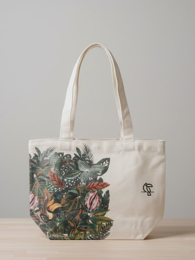 A white cotton canvas tote bag featuring a vibrant, artistic print of green tropical leaves across the bottom and side